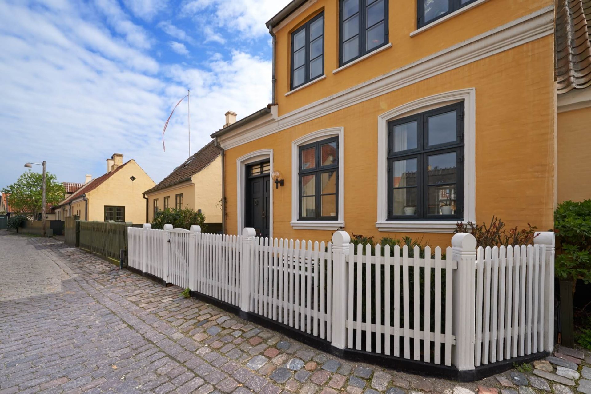 Längs en gata av kullersten står en villa målad i gult med svarta fönster och en grind i vitt trä